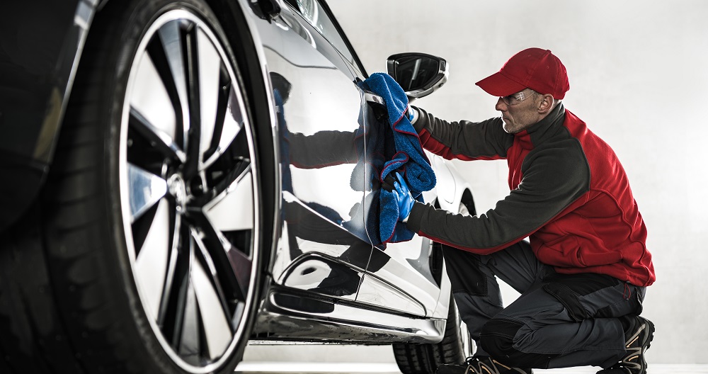 A person in a red cap and jacket is polishing the side of a car with a blue microfiber cloth in a well-lit space, showcasing expert mobile detailing services in LaPlace.
