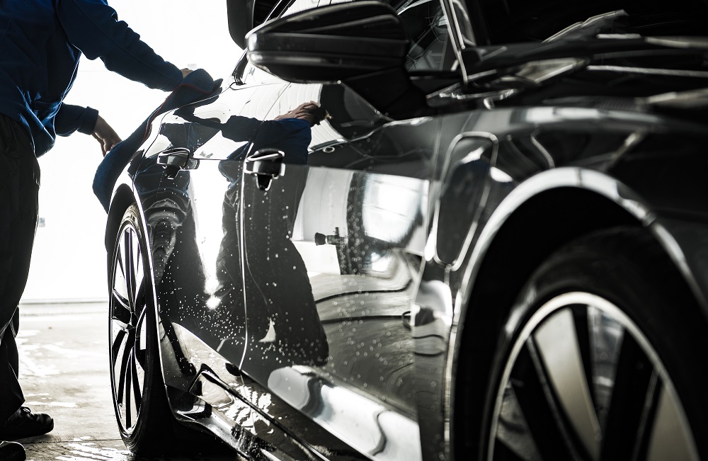 A person in blue overalls washes a black car covered in soapy water, its rear lights illuminated—showcasing the quality of Car Detailing Services in Madisonville.