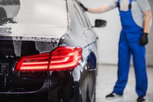 A person wearing a blue jacket is polishing the side of a shiny black car, showcasing expert Car Detailing Services in Madisonville with soapy water and reflections gleaming on the vehicle's surface.