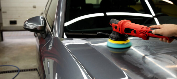 A person uses a red electric polisher with a foam pad to polish the hood of a gray car in a garage during a car detailing session.