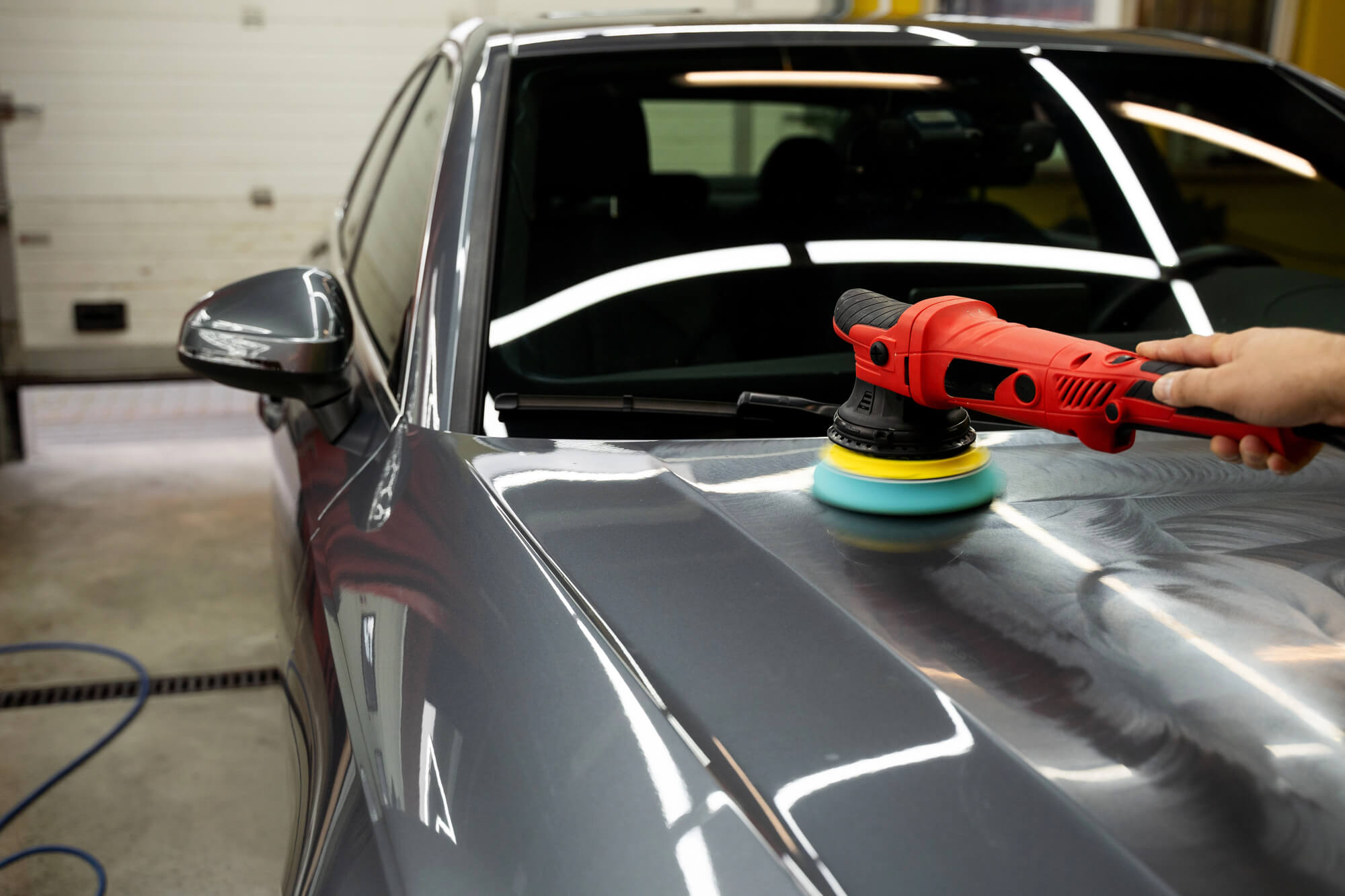 A person uses a red electric polisher with a foam pad to polish the hood of a gray car in a garage during a car detailing session.